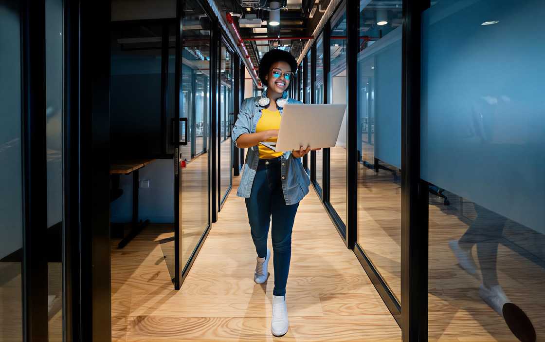 A woman walking through a modern office hallway holding a laptop.