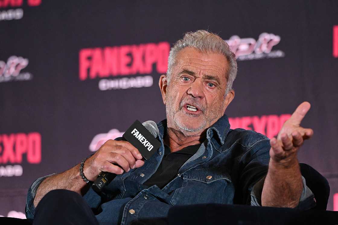 Mel Gibson wearing a denim jacket, speaking into a microphone and gesturing with his hand at a FanExpo panel.