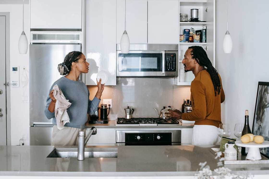 A couple argue in a modern kitchen while holding household items.