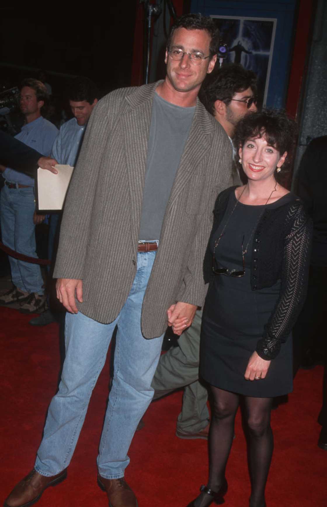 A tall man in a gray blazer and jeans stands beside a woman in a black knit dress on a red carpet.