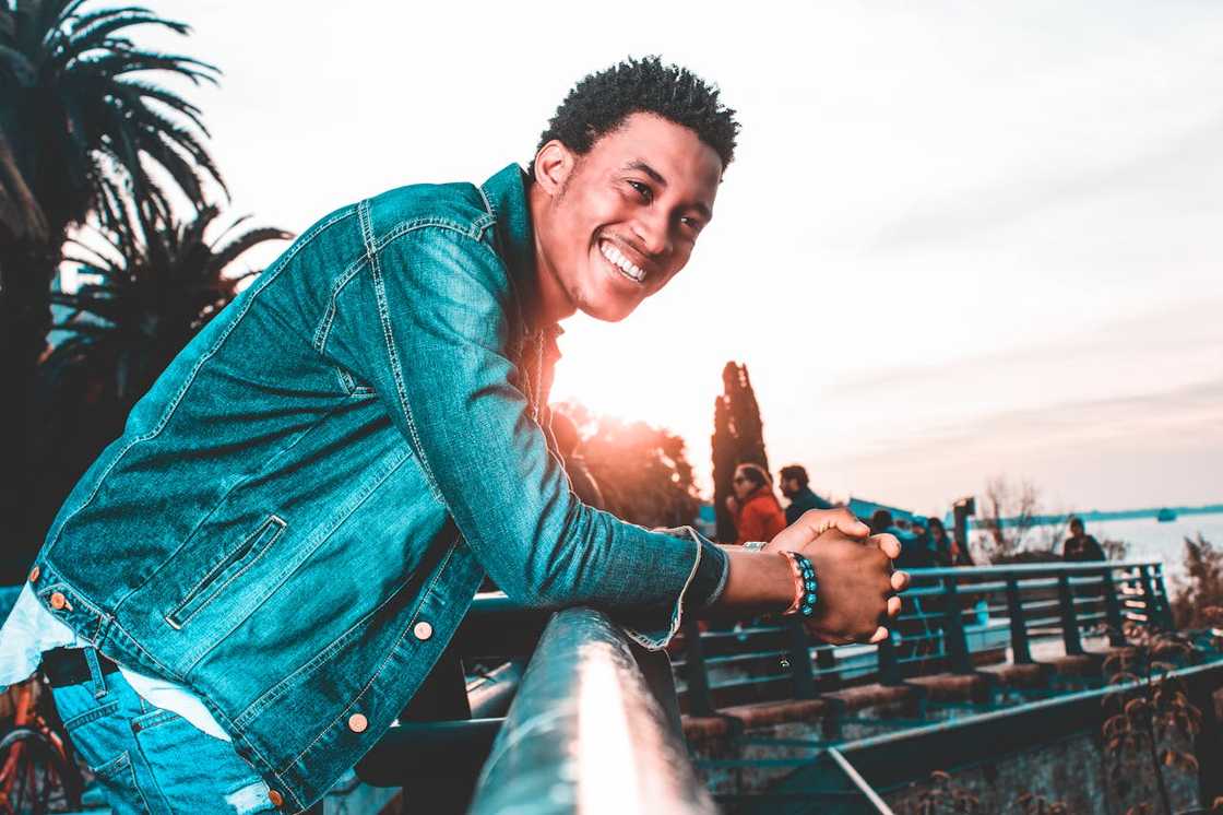 A person in a denim jacket smiles, with palm trees and water in the background.