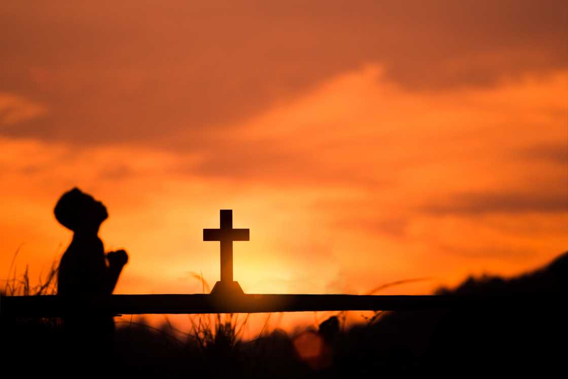 Silhouette of someone praying before a cross during sunset