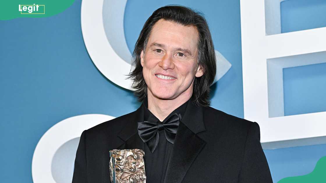 Jim Carrey in a black suit and bow tie holds a trophy in front of a blue Golden Globes backdrop.