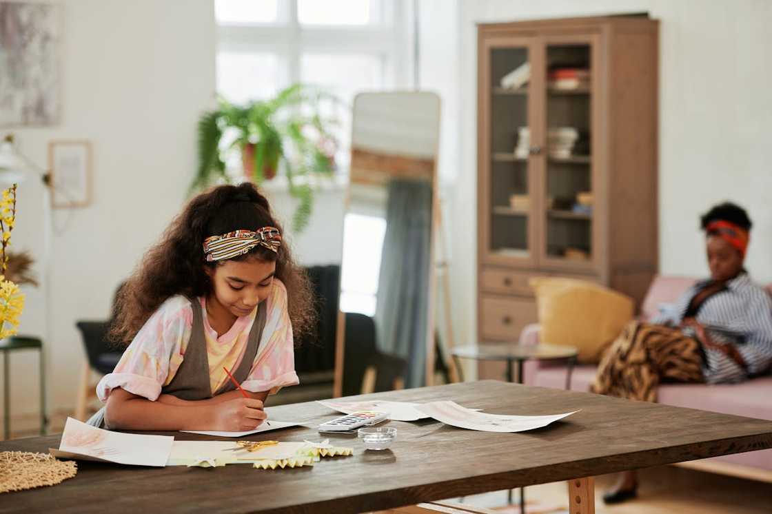 A girl draws at a table while an adult sits in the background. A girl draws at a table while an adult sits in the background.
