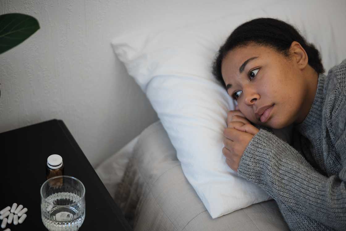 A sick woman lying on bed near bedside table with pills and glass of water