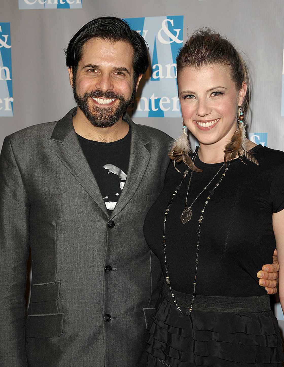 A bearded man in a gray blazer stands beside a smiling woman in a black top with feather earrings.