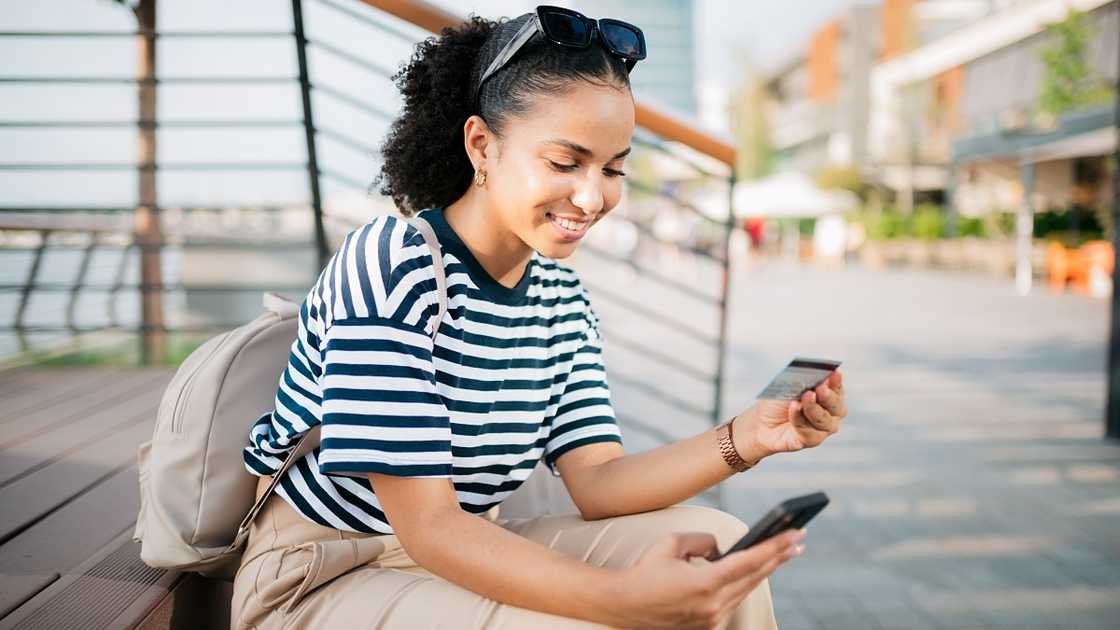 A woman is pictured sitted, checking her credit card and phone.