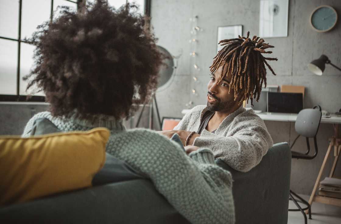 Two people sit on a couch in a cozy, sunlit room, engaged in conversation. Two people sit on a couch in a cozy, sunlit room, engaged in conversation.