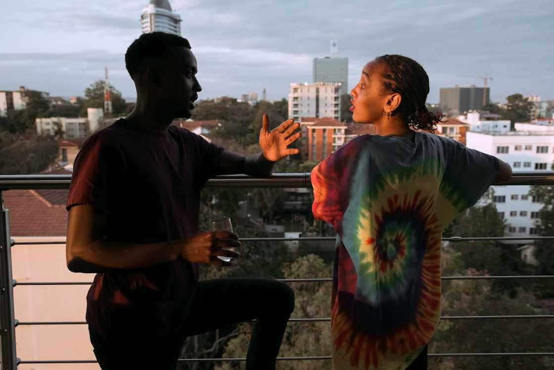 A man gestures while speaking to a woman on a balcony overlooking a city.