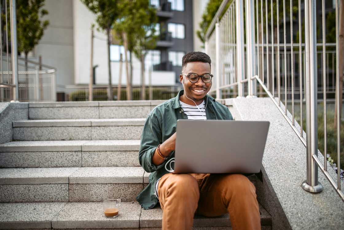 A guy sitting on the stairs in a front of the office building, working using laptop.