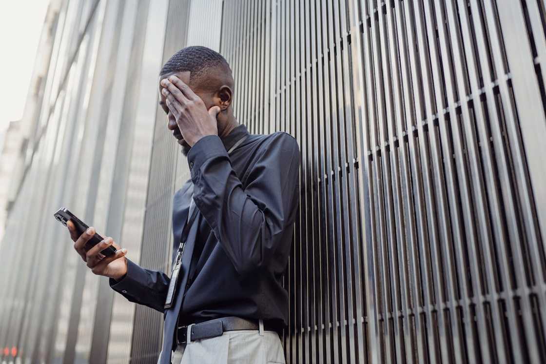 A man stands against a metal wall holding his phone and covering his face. A man stands against a metal wall holding his phone and covering his face.