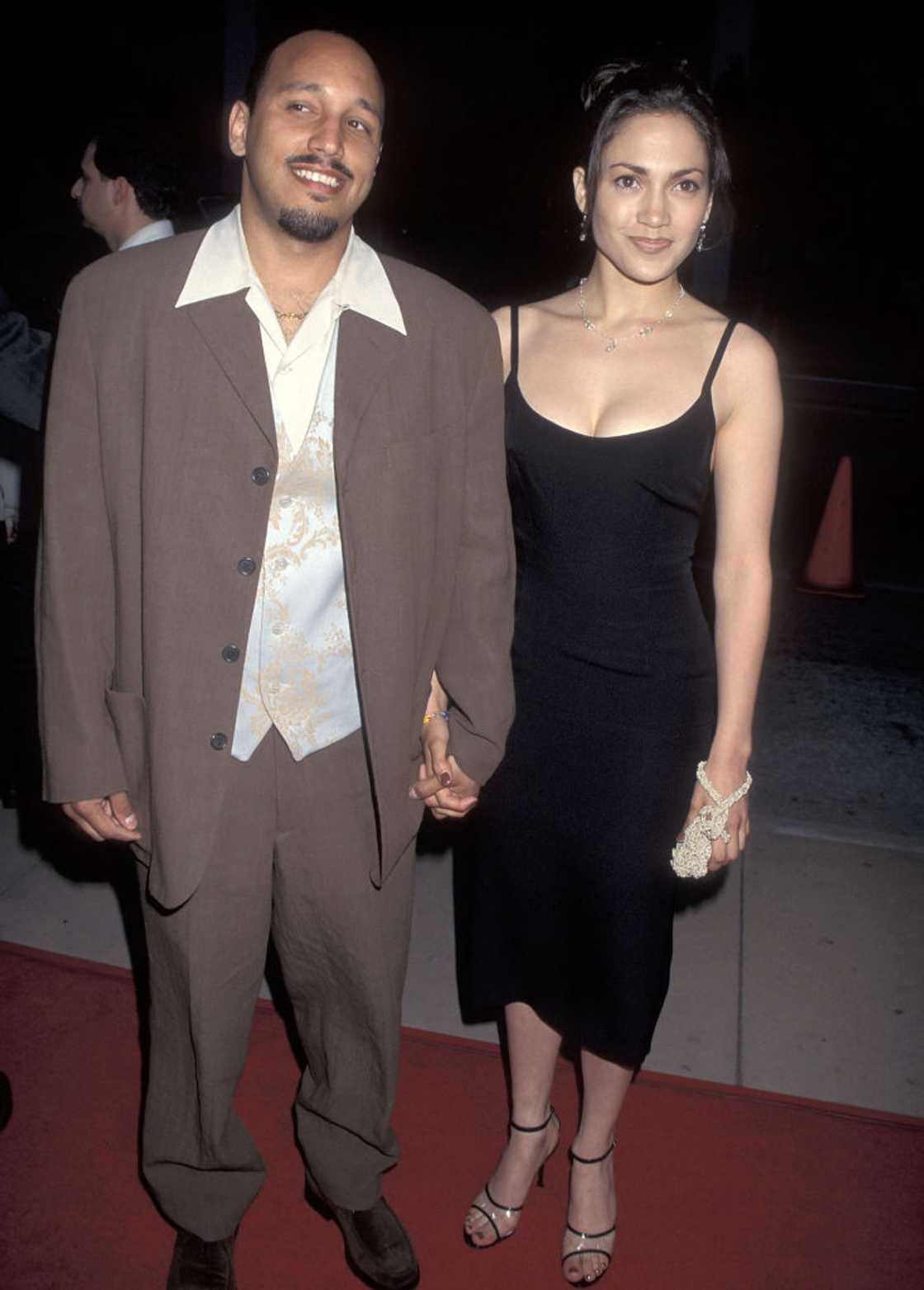 A man in a brown suit and a woman in a black slip dress smiling together on a red carpet. A man in a brown suit and a woman in a black slip dress smiling together on a red carpet.