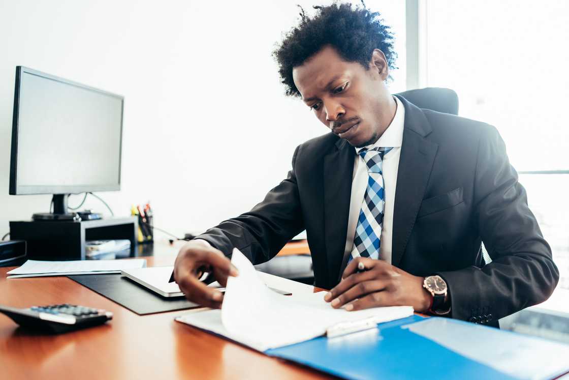 Man in a blue suit reviews documents at a desk in a sunlit office.