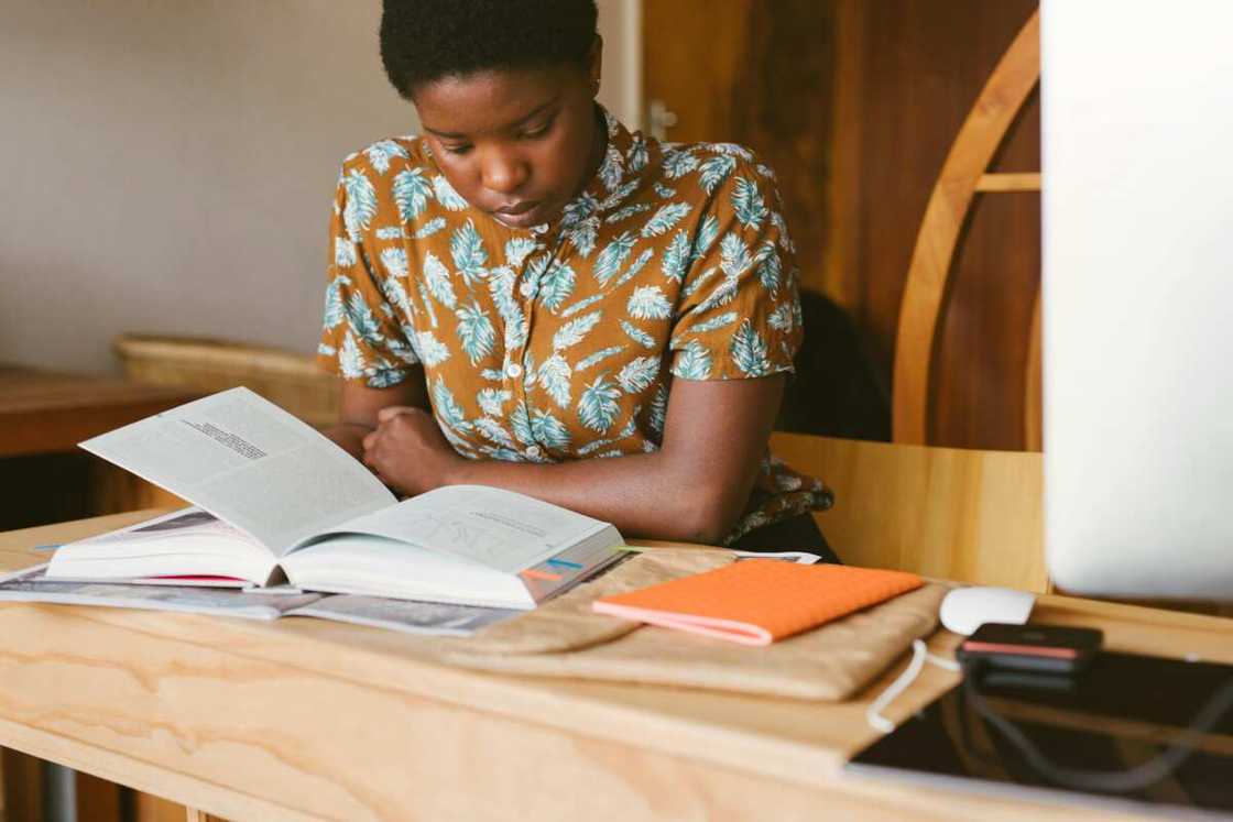 A young man in a floral top is reading a book on a desk