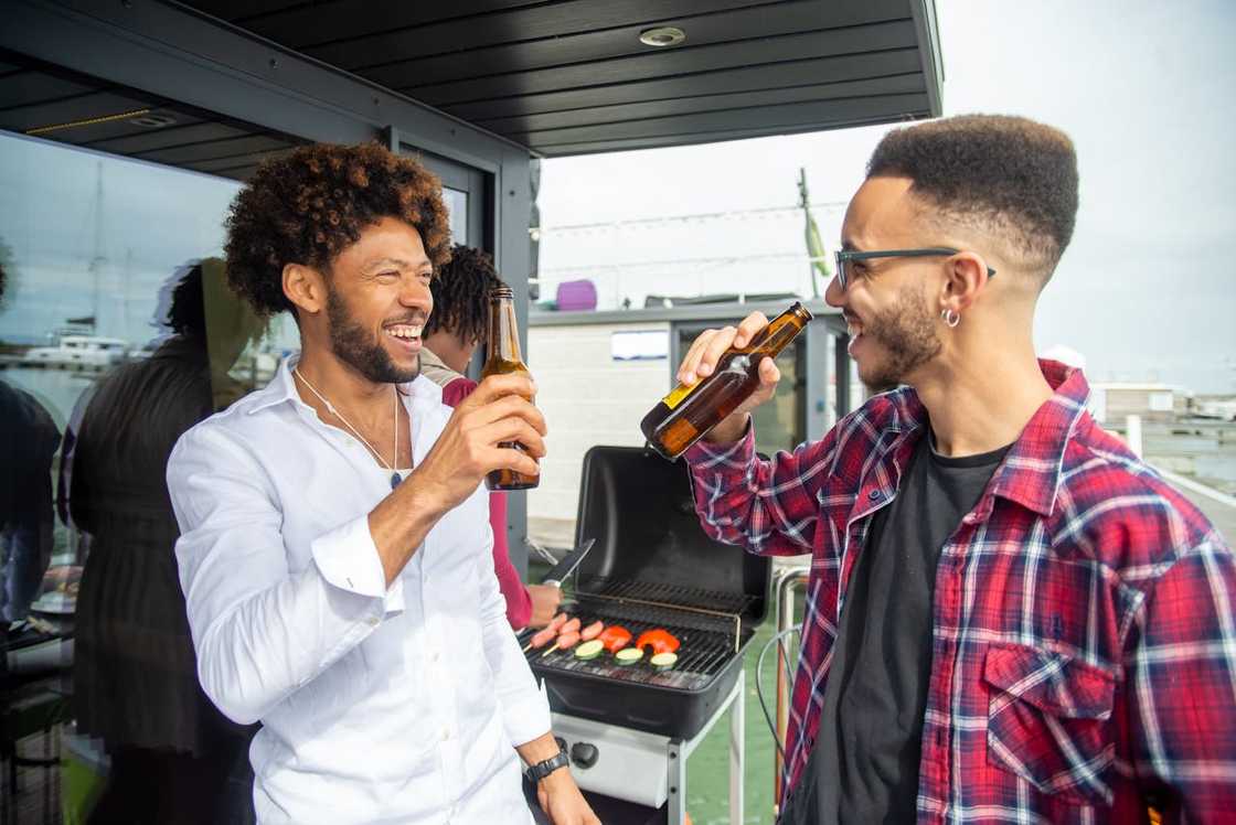 Two friends smiling and drinking beer near a barbecue grill.