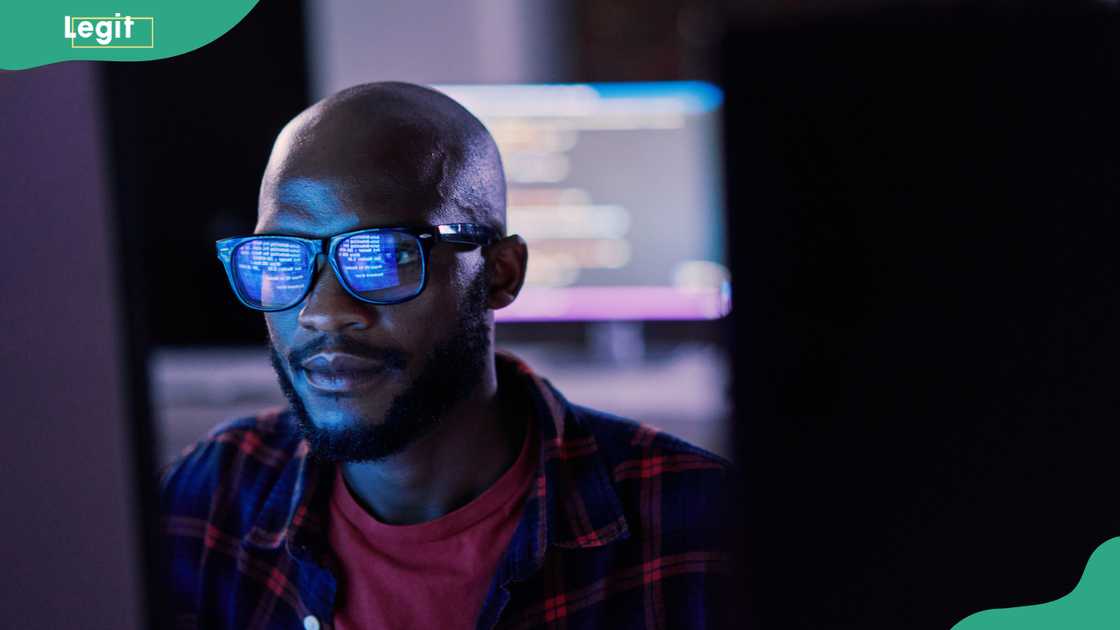 A bald man in a plaid shirt over a red t-shirt sits before glowing computer monitors, code reflected in his glasses.