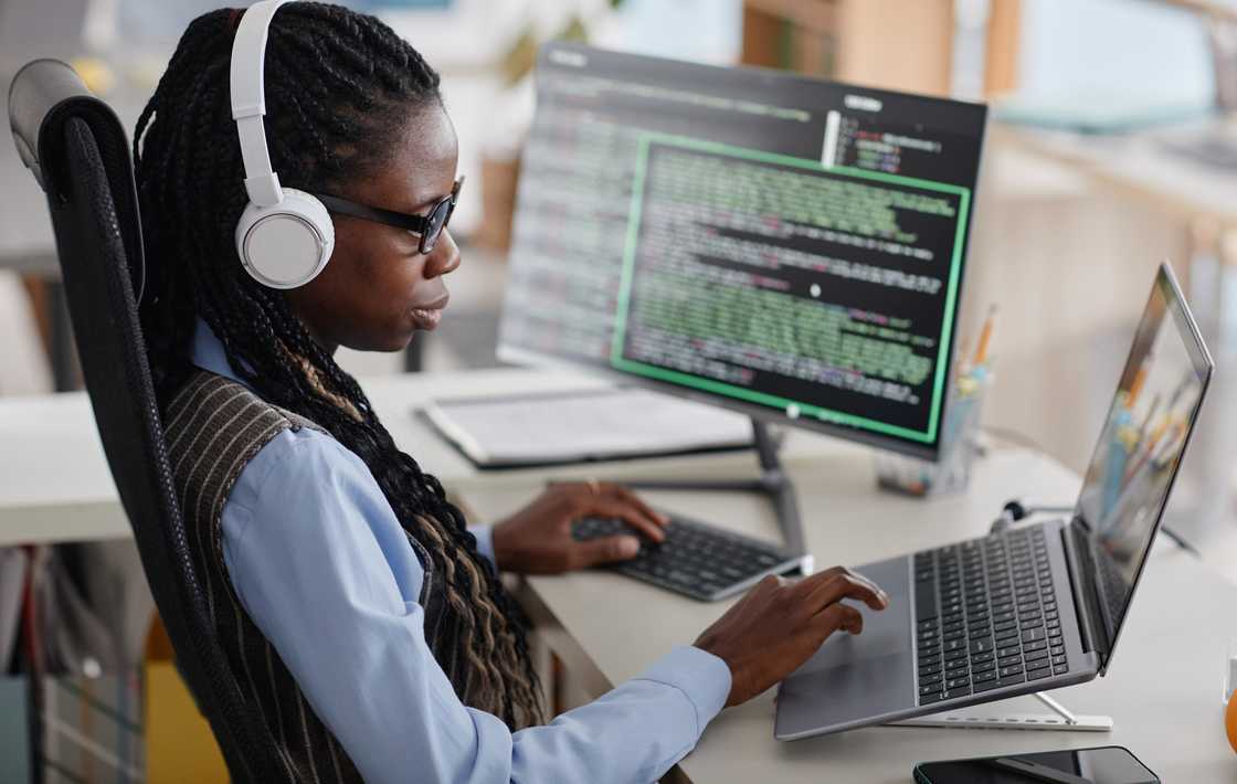 A female software engineer seats on a desk with multiple screens deeply engaged in coding while wearing headphones. A female software engineer seats on a desk with multiple screens deeply engaged in coding while wearing headphones.