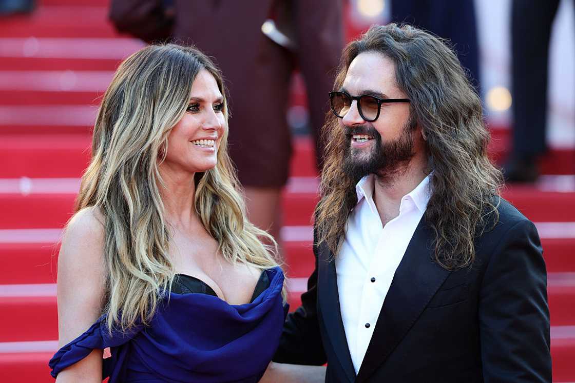 A woman with long, highlighted, wavy hair in a black and navy draped off-shoulder gown stands close to a man in sunglasses. A woman with long, highlighted, wavy hair in a black and navy draped off-shoulder gown stands close to a man in sunglasses.