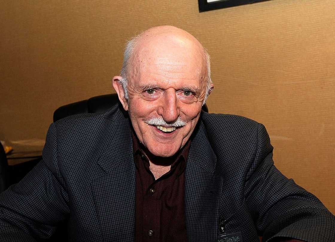 John Astin poses for a photo in a checked charcoal grey coat and a maroon shirt at the Parsippany Hilton