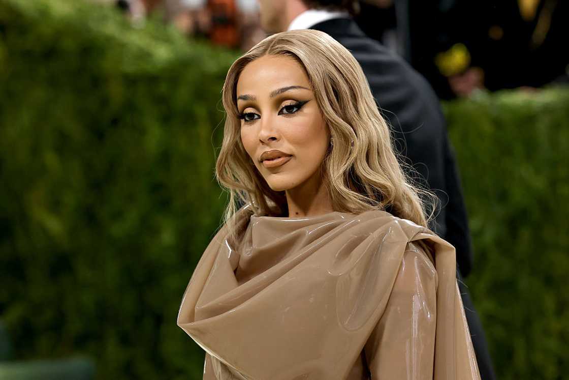 A woman with wavy blonde hair looks over her shoulder wearing a beige draped top against a green hedge backdrop. A woman with wavy blonde hair looks over her shoulder wearing a beige draped top against a green hedge backdrop.