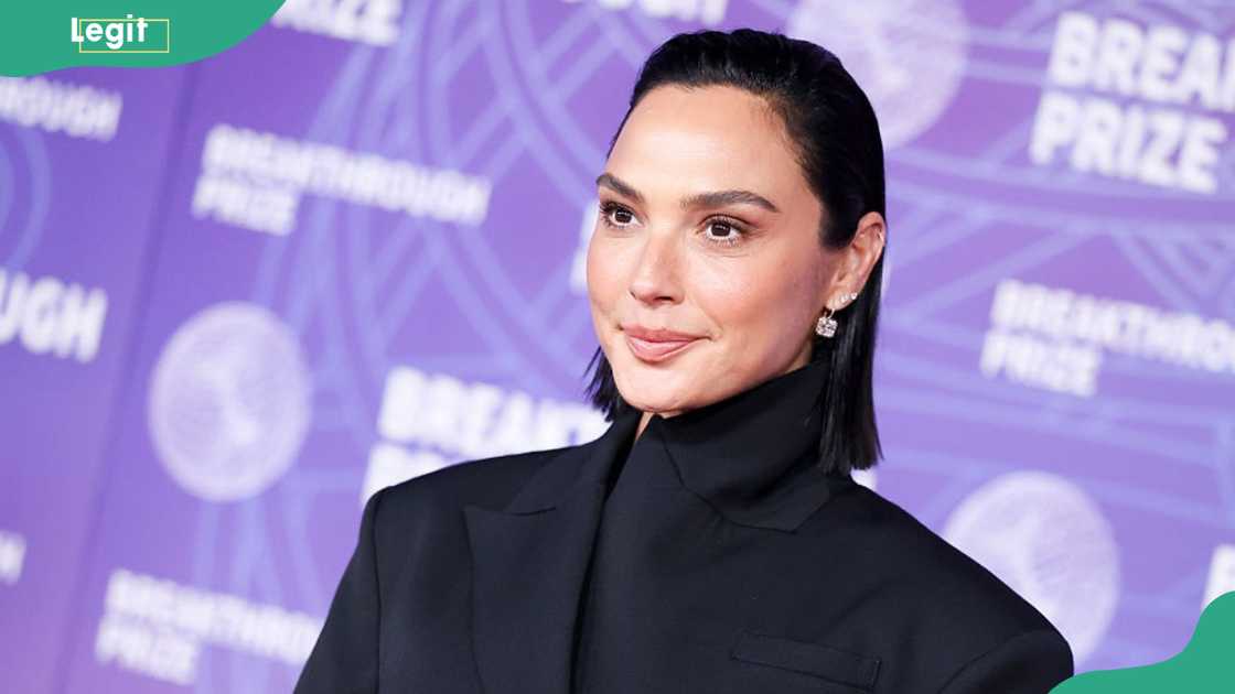 A dark-haired woman in an oversized black blazer looks off to the side at a purple step-and-repeat.
