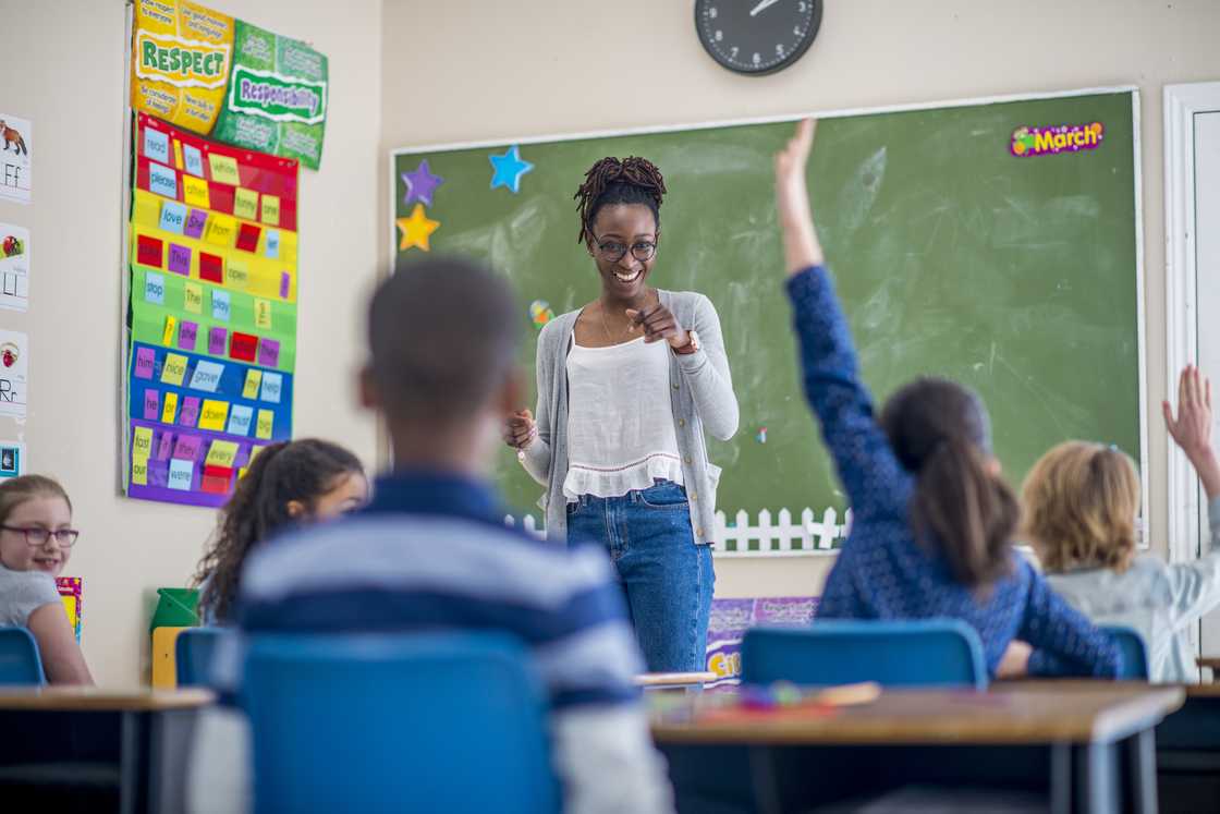 Teacher smiles and points toward students with raised hands in a colourful classroom.