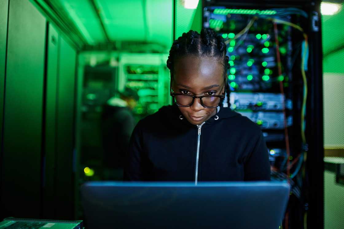 A computer programer using a laptop while standing in a dark server room A computer programer using a laptop while standing in a dark server room