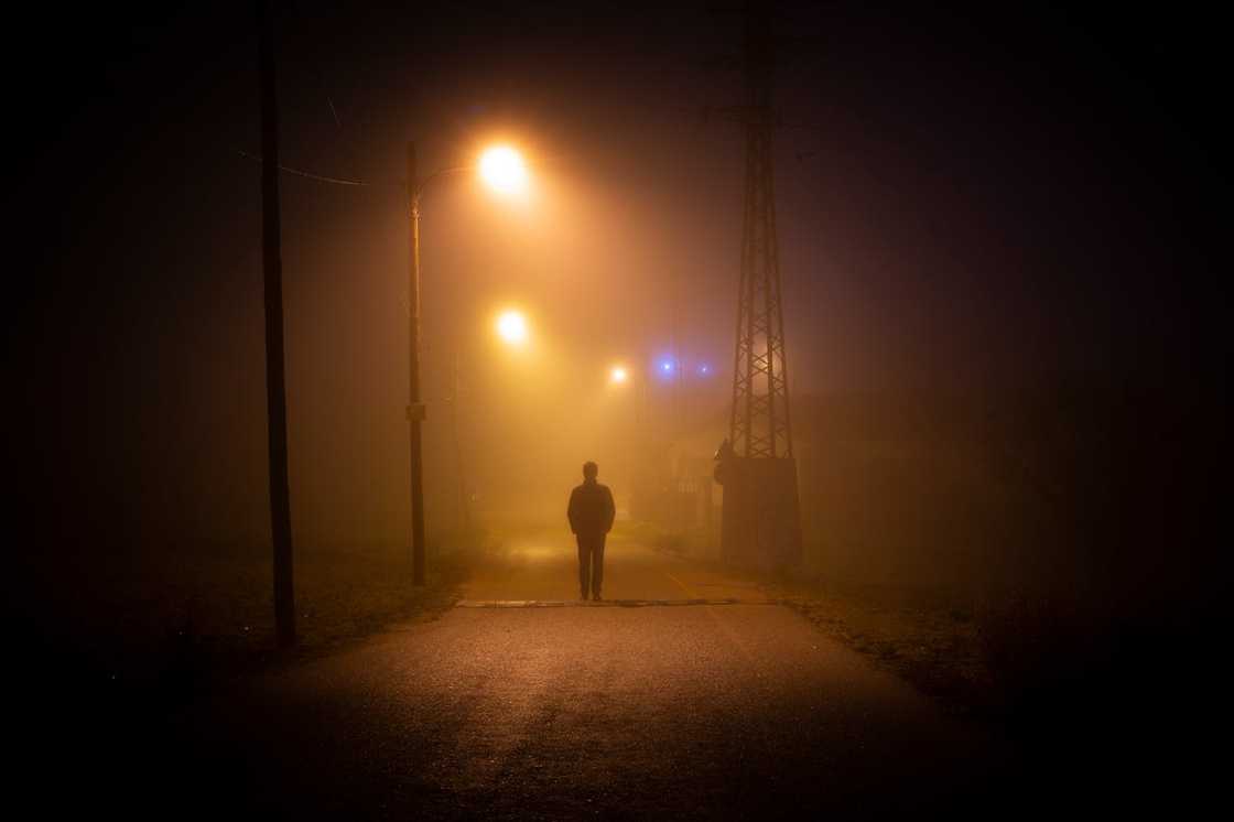A person walks alone down a foggy street at night under streetlights. A person walks alone down a foggy street at night under streetlights.