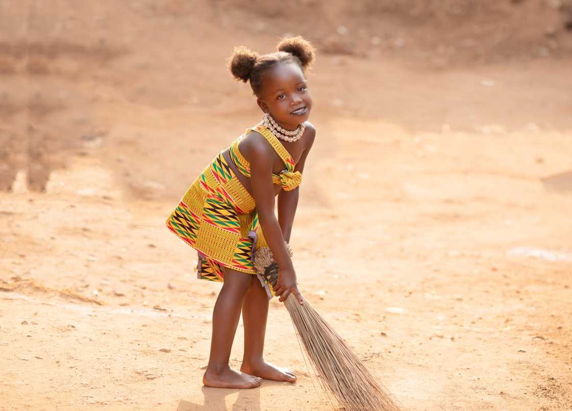 A young girl in a patterned dress holds a broom outdoors.