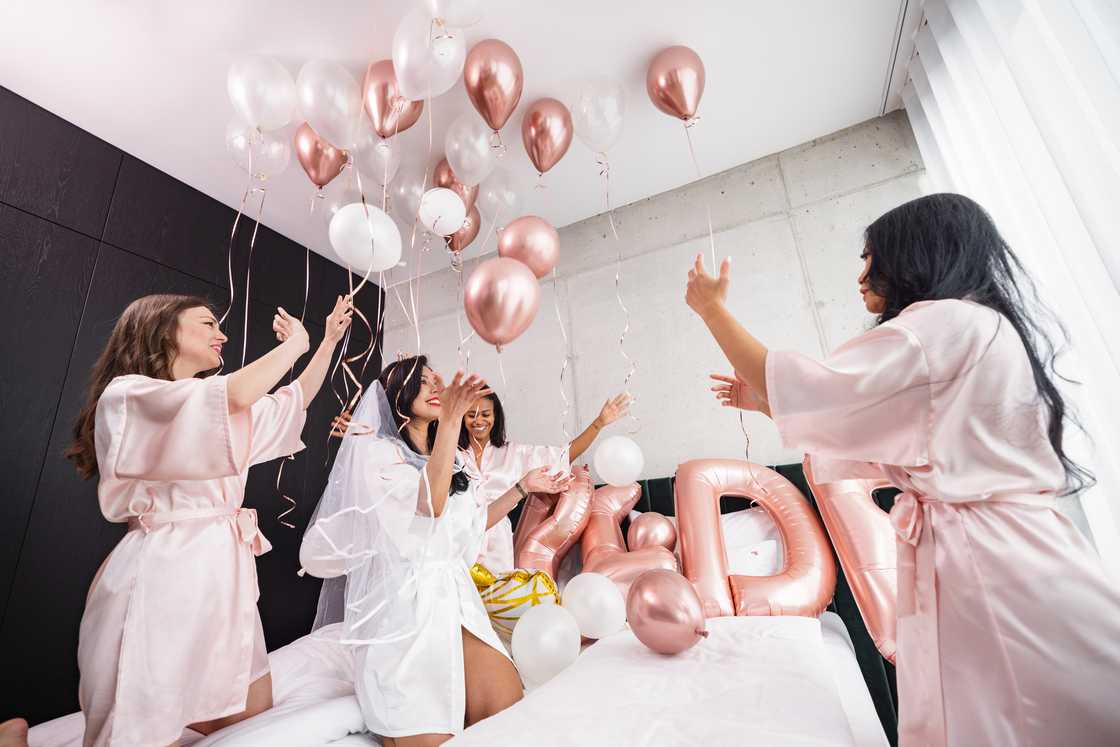 A group of friends jumping on the bed and playing with pink balloons during a bachelorette celebration