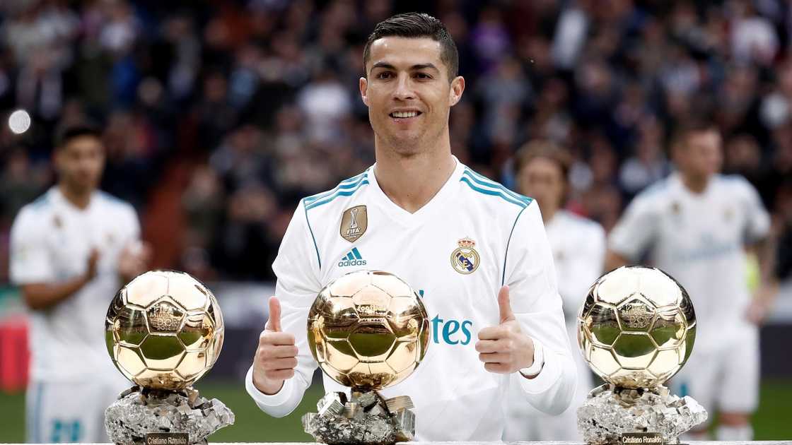 Cristiano Ronaldo poses standing with three of his five Ballon d'Or trophies on a football field.