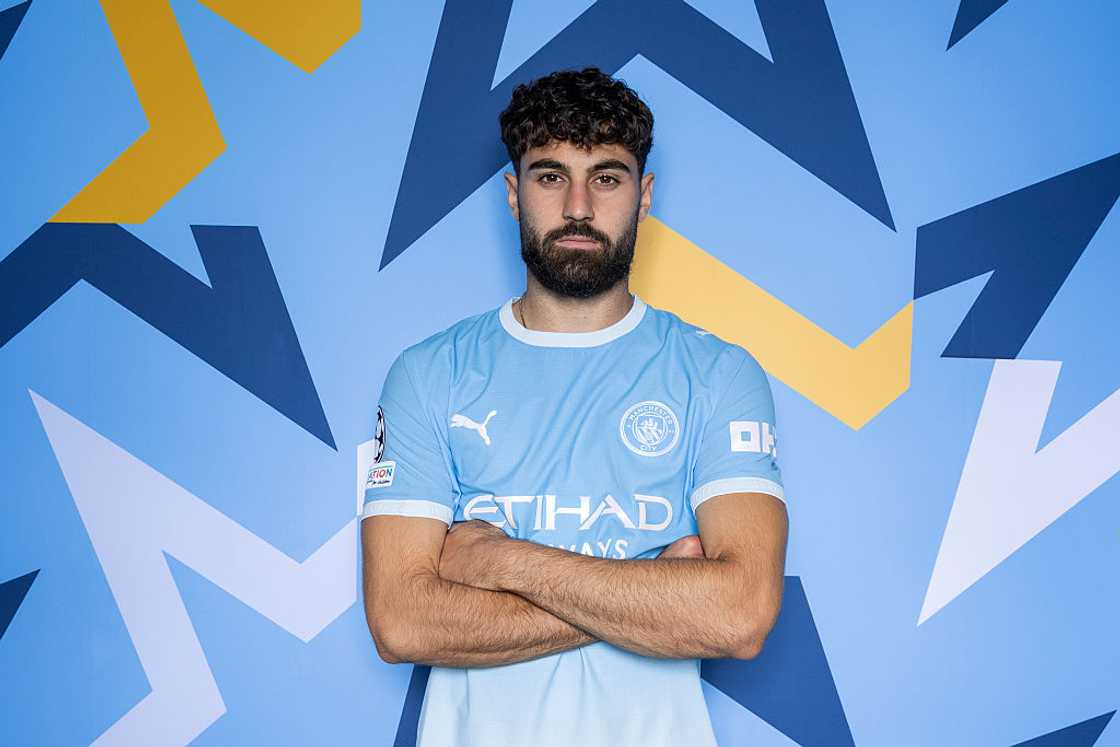 Joško Gvardiol, in the sky-blue Manchester City home kit, poses with arms folded against a blue patterned backdrop. Joško Gvardiol, in the sky-blue Manchester City home kit, poses with arms folded against a blue patterned backdrop.