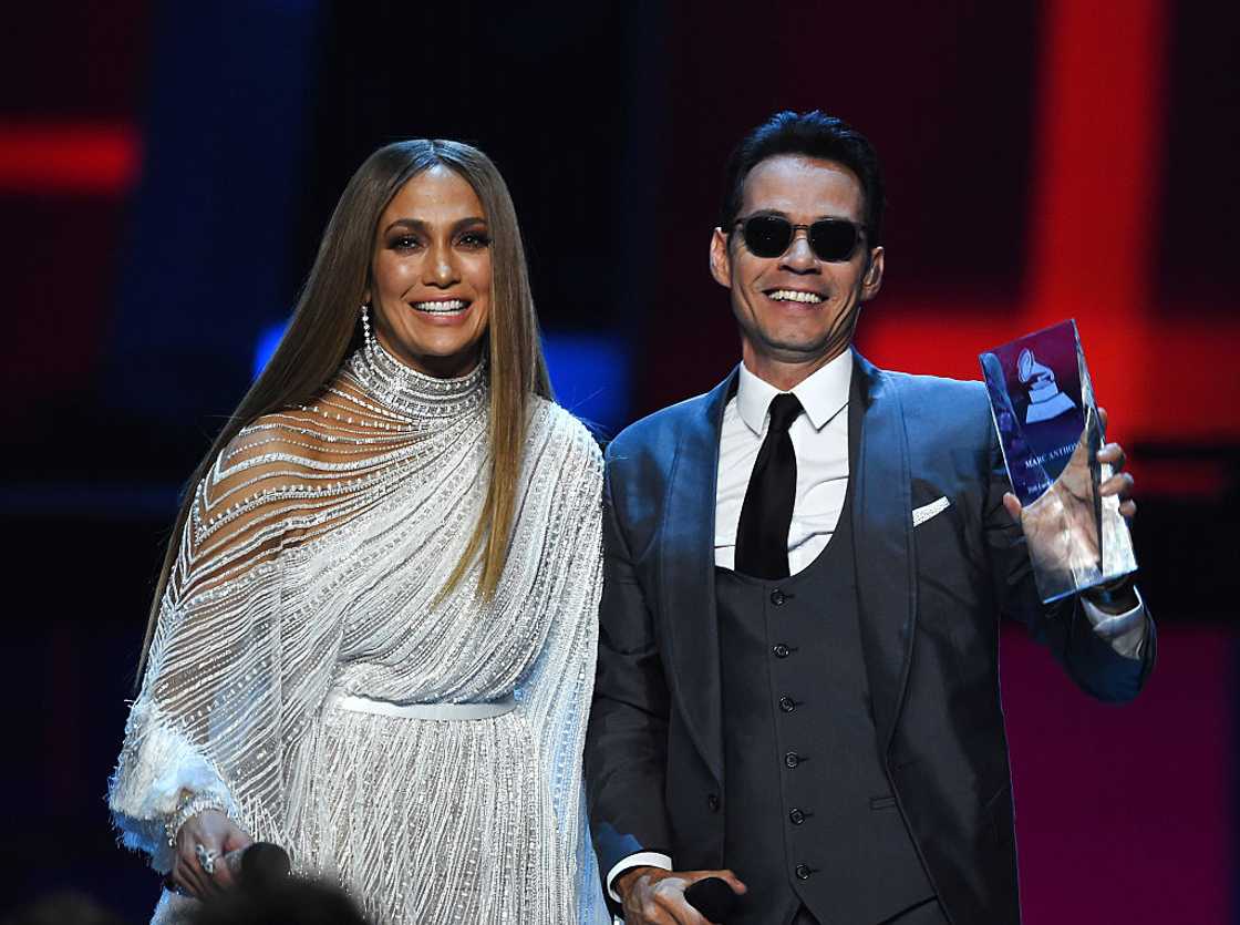 Jennifer Lopez in a white embellished gown smiling beside Marc Anthony holding a trophy. Jennifer Lopez in a white embellished gown smiling beside Marc Anthony holding a trophy.