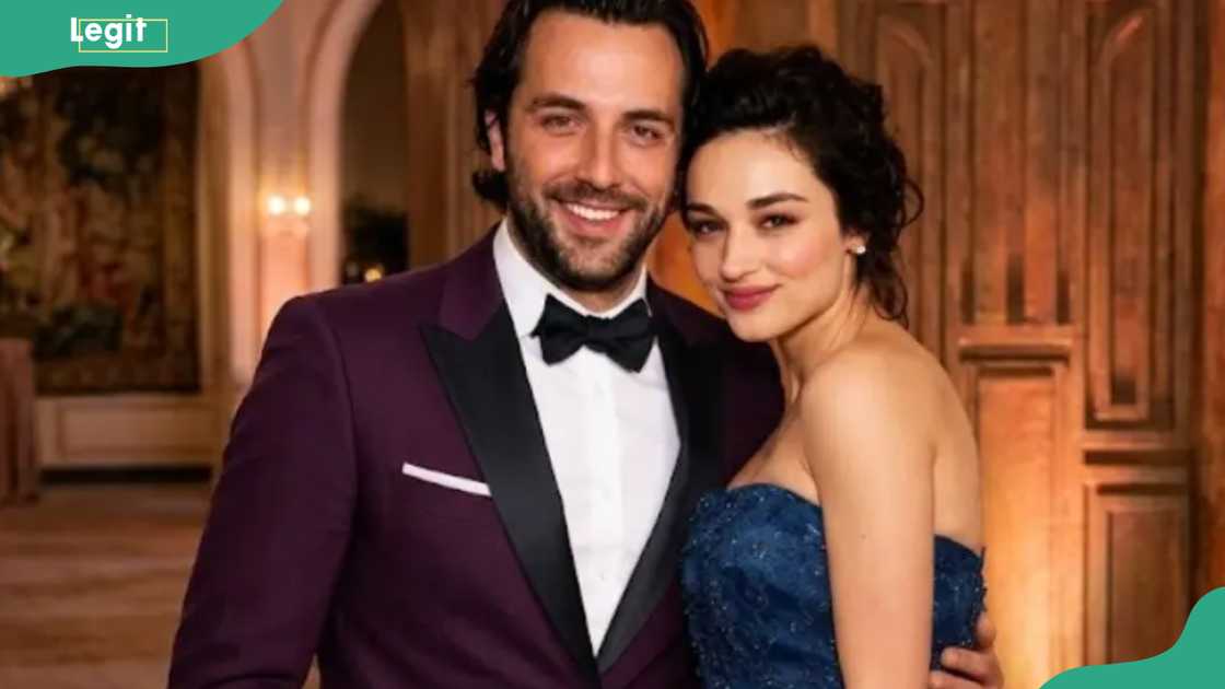 A bearded man in a maroon tuxedo and bow tie poses with a dark-haired woman in a sparkly blue strapless dress.