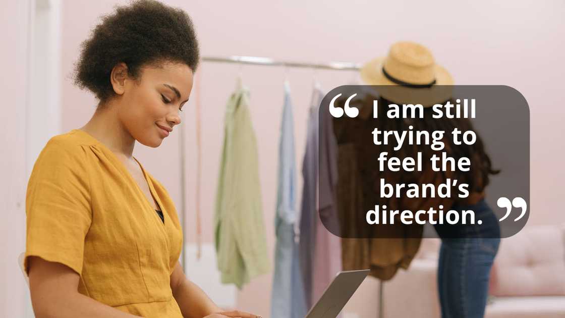 A woman uses a laptop while another sorts clothes on a rack.