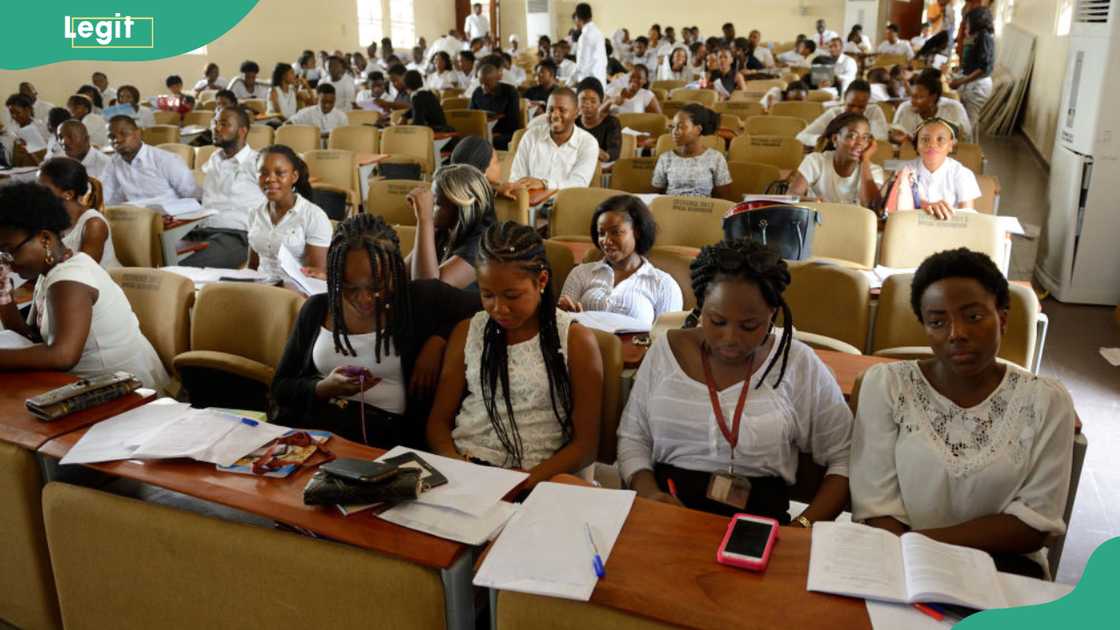 Rows of students sit at desks with notebooks and papers in a crowded lecture hall, with several in the foreground writing and reading.