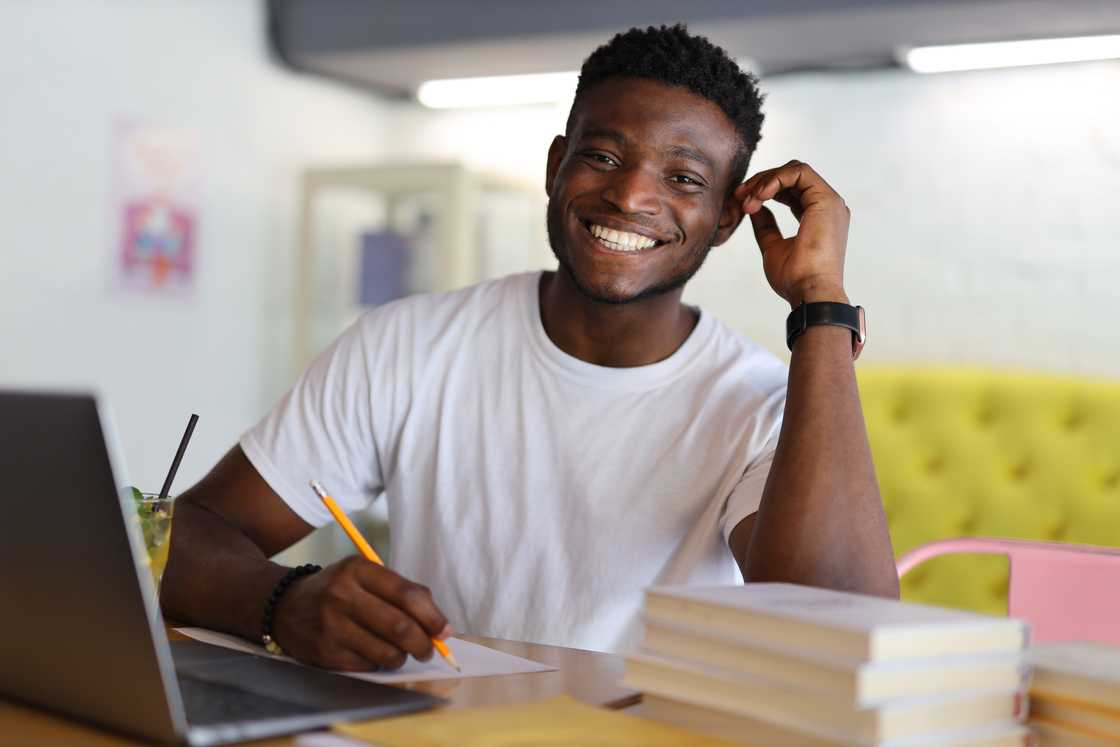 A serious and cheerful young man, immersed in education, studying at home with books, a laptop, and a notebook.