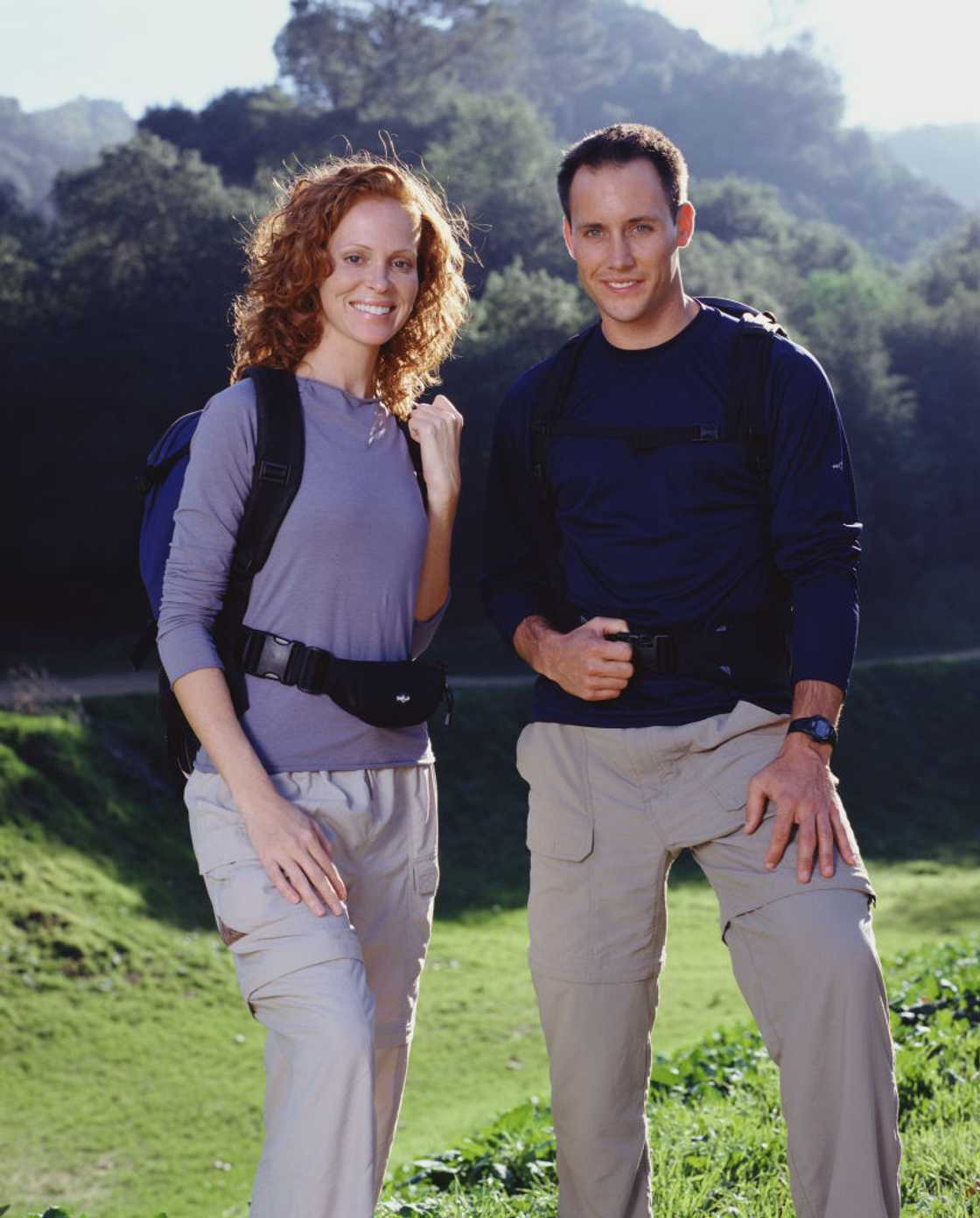 A woman with curly red hair and a man in a navy top stand together on a grassy hillside, both wearing backpacks and hip bags.