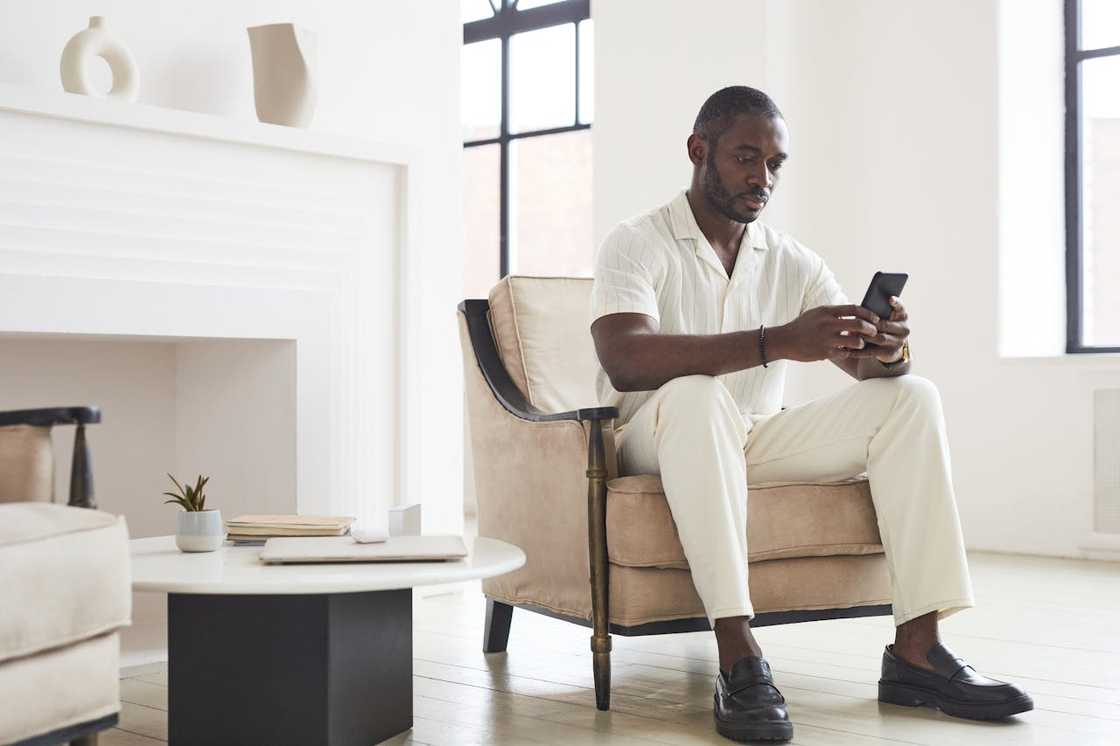 A man sits alone in a chair looking at his phone in a bright living room.
