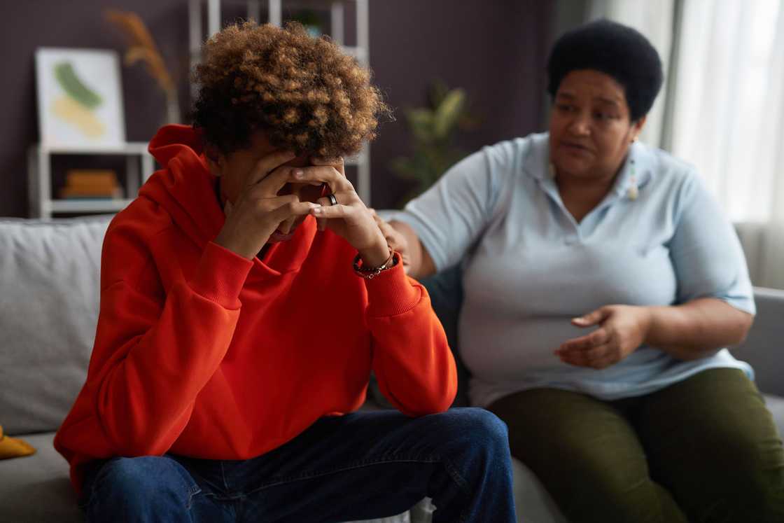 An elderly woman confronts a seated young man in the living room.