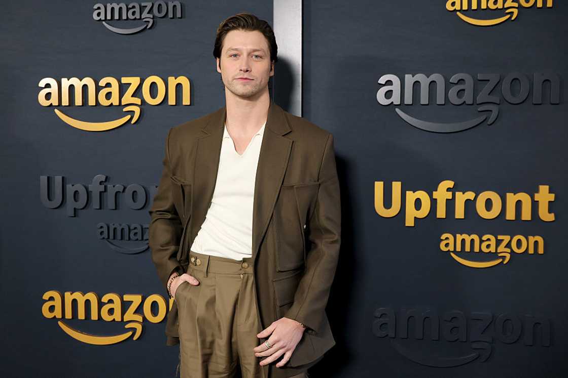 man in a brown blazer and cream shirt stands with hand in pocket in front of an Amazon Upfront backdrop. man in a brown blazer and cream shirt stands with hand in pocket in front of an Amazon Upfront backdrop.