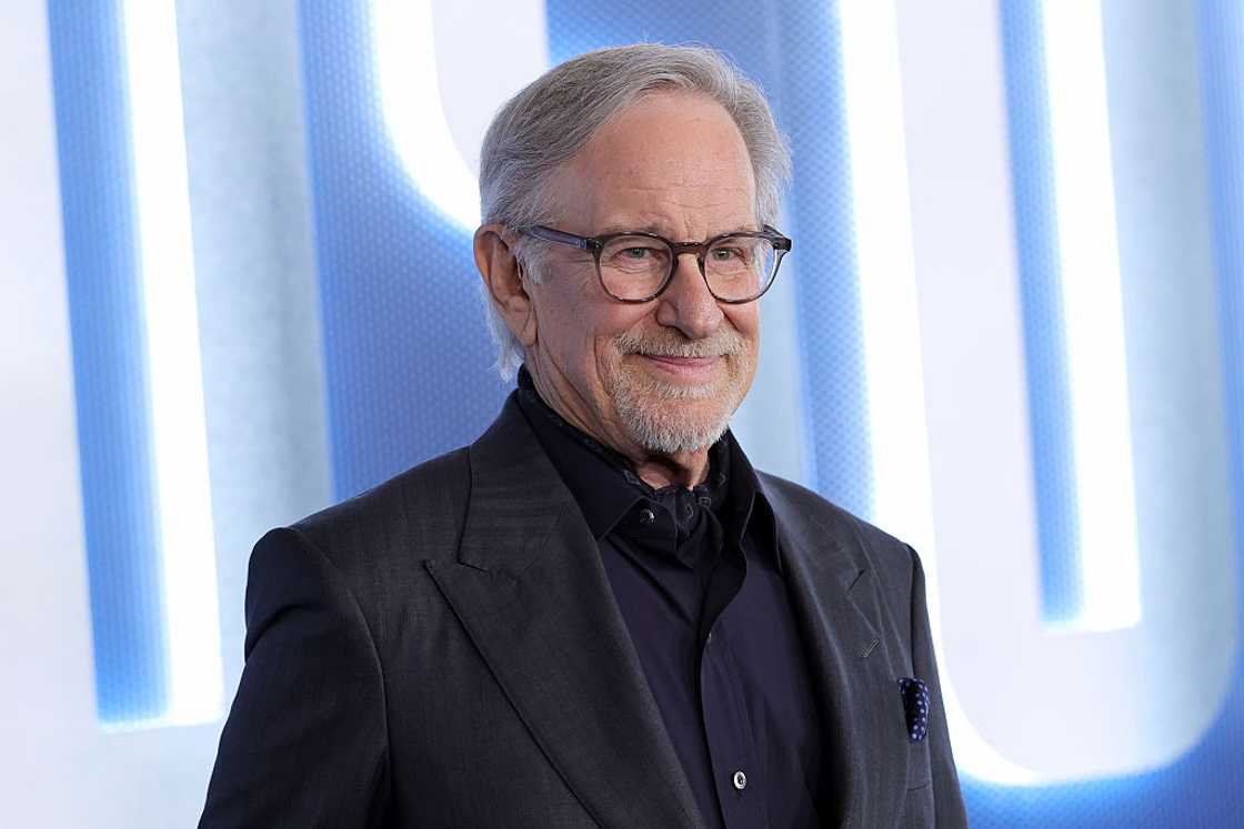 Steven Spielberg wearing glasses and a black suit with a black shirt, smiling against a blue and white backdrop.