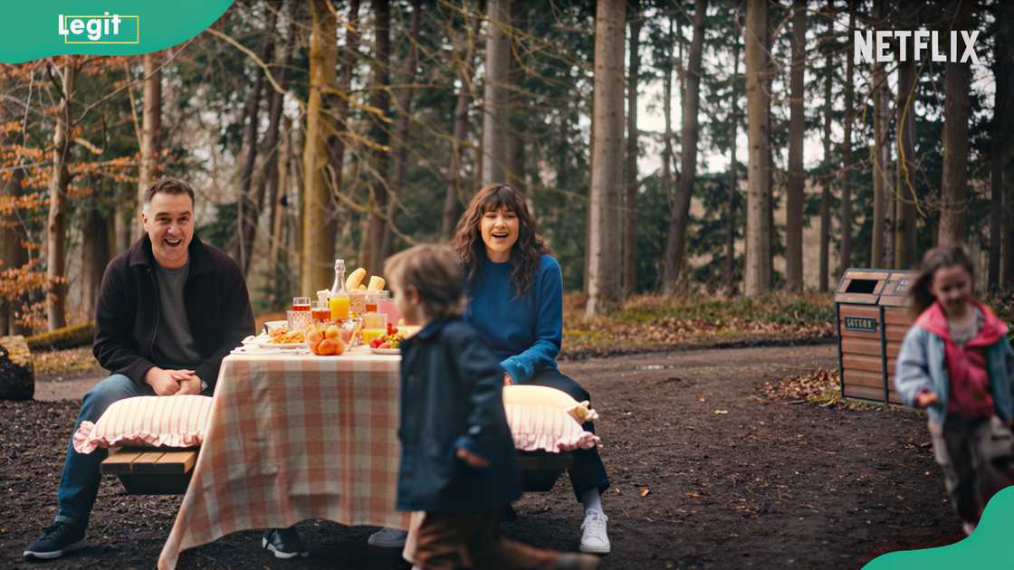 Two adults sit at a picnic table in a forest while two kids run past