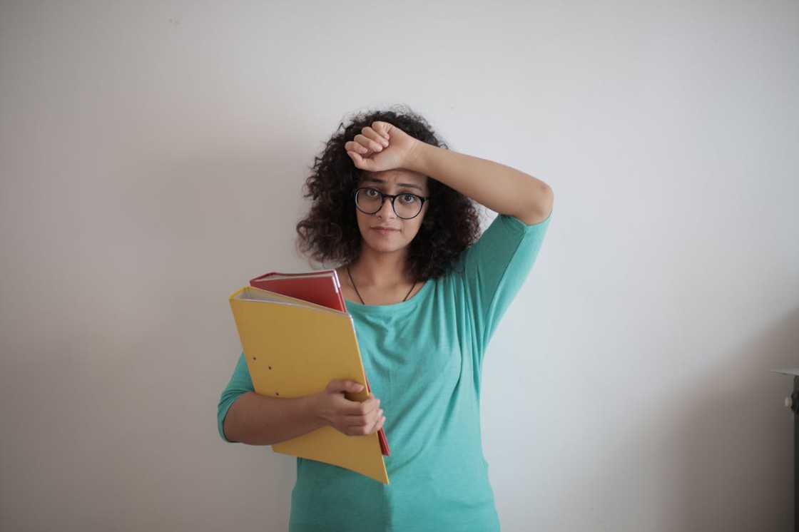 A woman holding folders, raising her arm to her forehead in a stressed gesture.