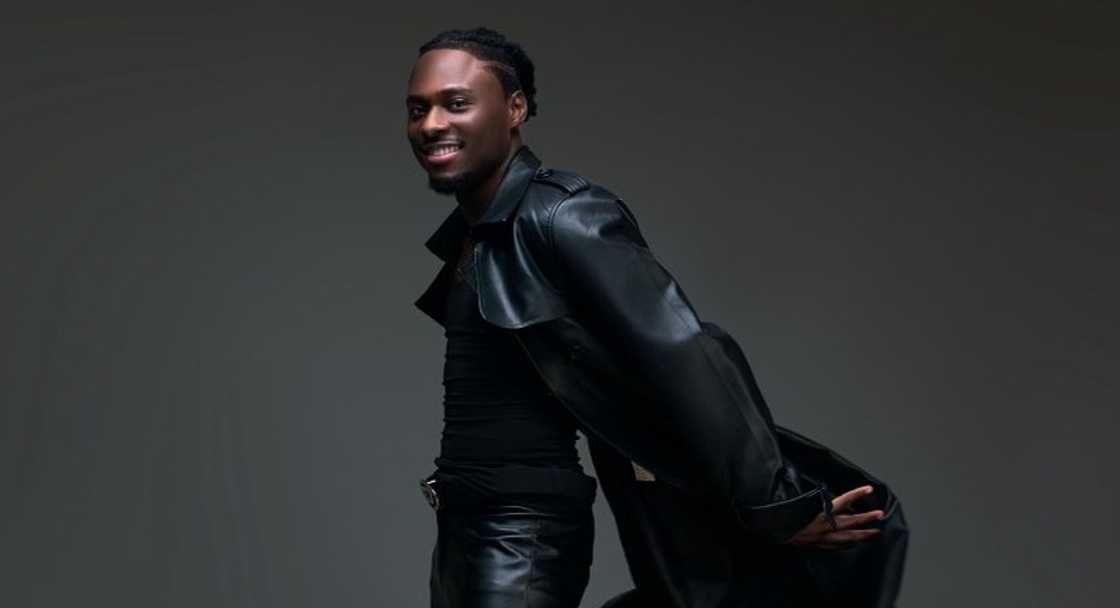Faith from BBNaija flicks his black jacket during a photoshoot against a grey background.