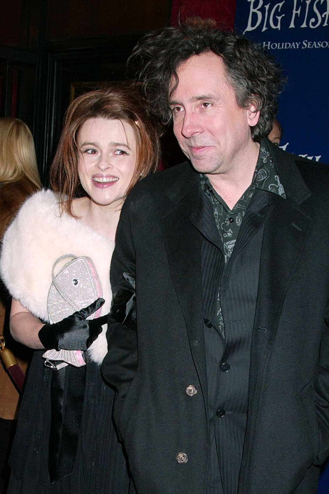 A man in a dark patterned shirt stands with a woman in a white fur wrap holding a small bag.