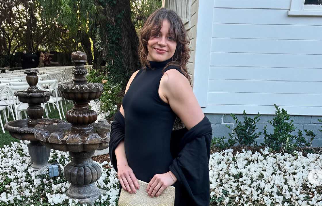 Ashley Basset pictured in a black dress and holding a gold clutch poses in an outdoors garden setting