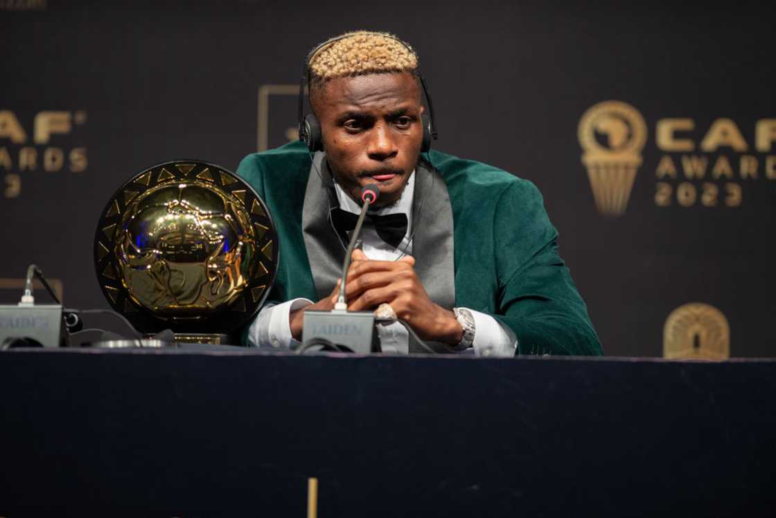 Victor Osimhen in a green velvet tuxedo jacket and bow tie, speaking into a microphone