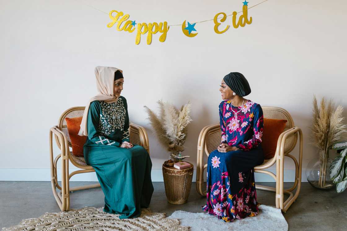 Two women in hijabs are sitting in front of a wall with an Eid decoration