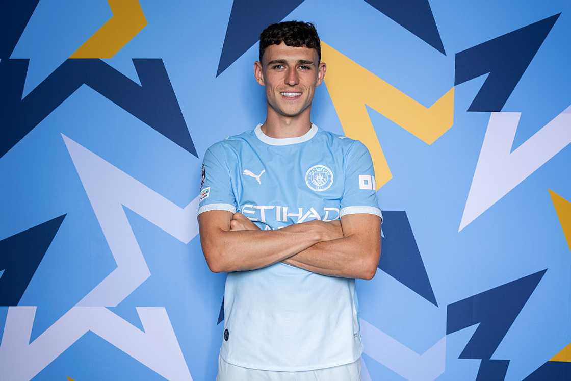 Phil Foden, in the sky-blue Manchester City home kit, poses with arms folded against a blue patterned backdrop. Phil Foden, in the sky-blue Manchester City home kit, poses with arms folded against a blue patterned backdrop.